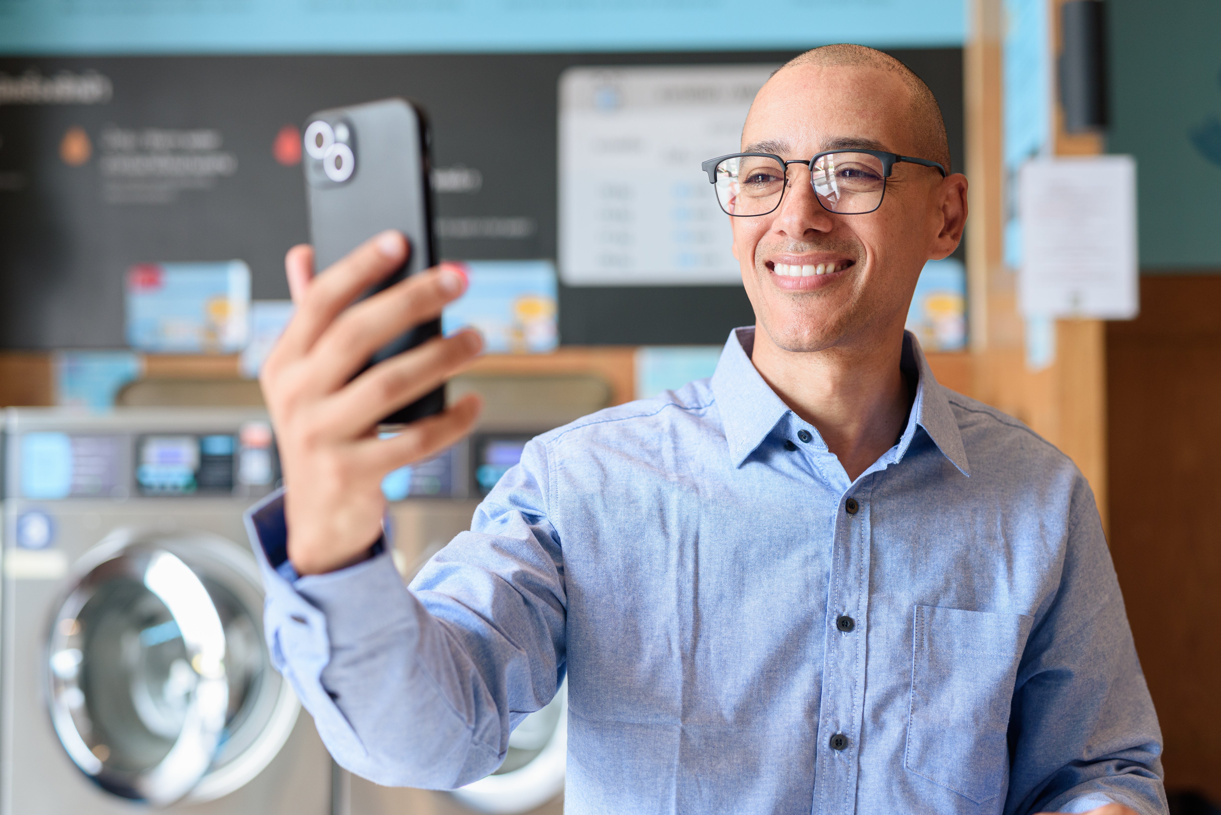 Man with glasses taking a selfie in a laundry room using a smartphone.